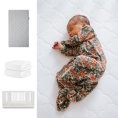 A baby sleeps on a white Newton Baby Nest Convertible Crib with the Original Mattress, alongside images of a gray mattress and two folded Breathable Organic Cotton Sheets. 111-8420-WHT-1000-GRY-WNW