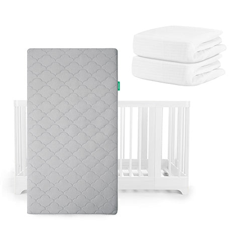 A gray scalloped Newton Baby Original Mattress stands before a white Nest Convertible Crib, with two folded white sheets stacked at its upper right.