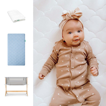 A baby in a brown polka-dot outfit with a matching headband lies on a Newton Baby Bundle 6 BFCM white quilted mattress. To the left are a folded white sheet, a light blue quilted mattress pad, and a wooden bassinet. 1000-blu-8400-2000