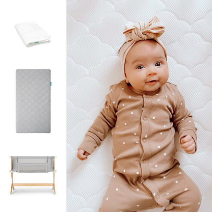 A baby in a brown polka dot onesie and striped headband lies on a Newton Baby Bundle 6 BFCM white quilted mattress, with a white pillow, gray mattress, and wooden-frame bassinet shown to the left. 1000-gry-8400-2000