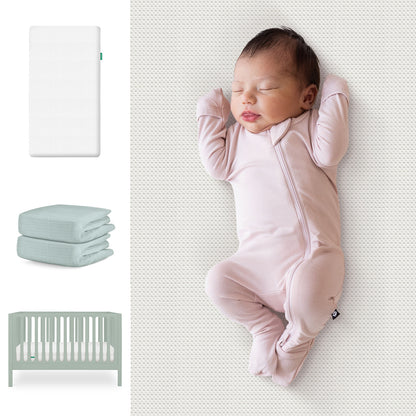 A baby in a light pink onesie sleeps on a light surface beside the Newton Baby Galileo Crib, Essential Crib Mattress, and Sheets Bundle. The crib, mattress, and two folded sheets are pictured to the left. 111-8420-SGE-1300-WHT-SNS