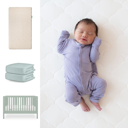 A baby in a lavender onesie sleeps on the Newton Baby Galileo Crib with the Original Crib Mattress and organic cotton sheets. Nearby are a beige mattress, folded light blue sheets, and the light green 3-in-1 Galileo Crib. 111-8420-SGE-1000-SAN-SNS
