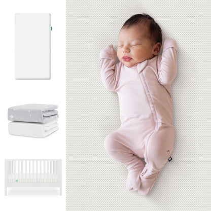 A sleeping baby in a light pink onesie lies on a white surface. To the left are the Newton Baby Galileo Crib, Essential Crib Mattress, and Sheets Bundle. 111-8420-WHT-1300-WHT-SGW