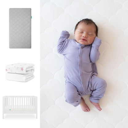 A baby in a purple onesie sleeps on a Newton Baby Original Crib Mattress. Nearby are the Galileo Crib, gray quilted sheets, and a pink star blanket—all from the Newton Baby Galileo Crib, Original Crib Mattress & Sheets collection. 111-8420-WHT-1000-GRY-PSW
