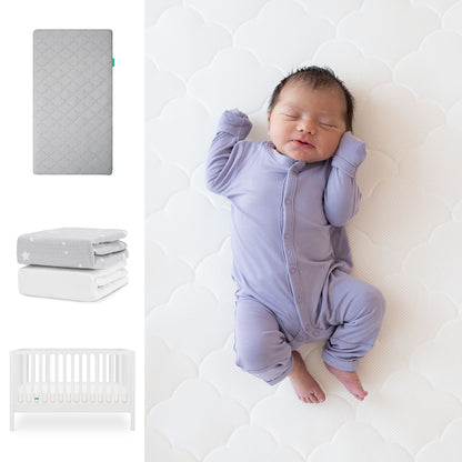 A baby in a lavender onesie sleeps on the Newton Baby Original Crib Mattress with a breathable organic cotton sheet. Nearby are the Galileo Crib, a gray mattress, and two folded blankets—one gray with stars, one white. 111-8420-WHT-1000-GRY-SGW
