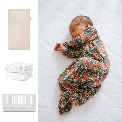 A baby in a floral swaddle lies on a white Newton Baby Nest Convertible Crib with the Original Mattress and breathable organic cotton sheets. Nearby are folded star-printed blankets, showcasing the Newton Baby set. 111-8420-WHT-1000-SAN-PSW