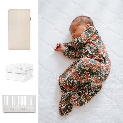 A newborn sleeps on Newton Baby’s Nest Convertible Crib with the Original Mattress and breathable organic cotton sheets; nearby, a floral knotted gown and a folded star-patterned blanket are also shown. 111-8420-WHT-1000-SAN-SDW