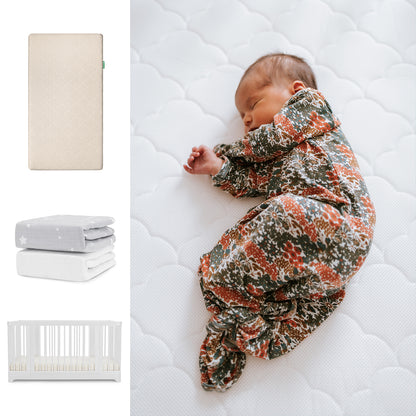 A newborn in a floral romper sleeps on the Newton Baby Nest Convertible Crib with Original Mattress. Nearby are grey and white Breathable Organic Cotton Sheets, alongside a cozy white crib from Newton Baby. 111-8420-WHT-1000-SAN-SGW