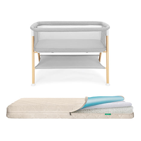 The Newton Baby Essential Bassinet + Waterproof Mattress features light wood legs, grey fabric sides, and a breathable mattress with its top layer partially unzipped to show the multiple internal layers.