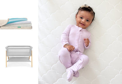 A smiling baby in a light purple onesie lies on a white, breathable Newton Baby Waterproof Mattress. Beside them is the Essential Bassinet + Waterproof Mattress in gray with wooden legs, shown with layered mattress details.