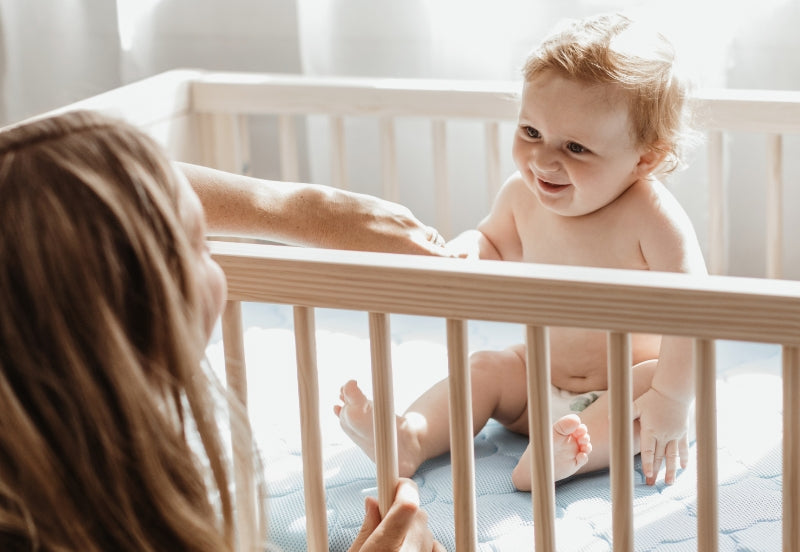 A joyful baby sits in a white crib, smiling and reaching out to a woman leaning over the railing.