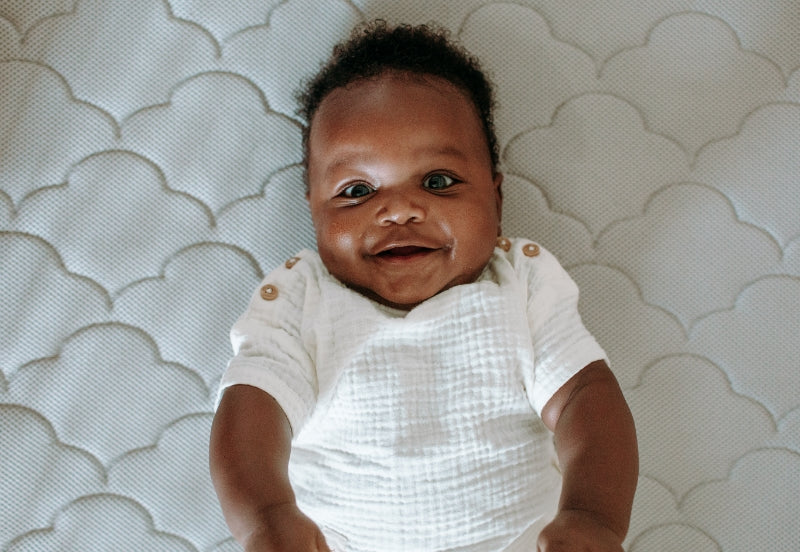 A cheerful baby laying on a gray, cloud-patterned mattress, smiling at the camera.