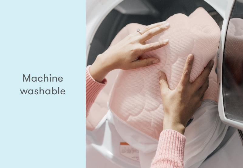 Hands placing a machine washable, pink baby mattress cover into a washing machine with the text "Machine washable."