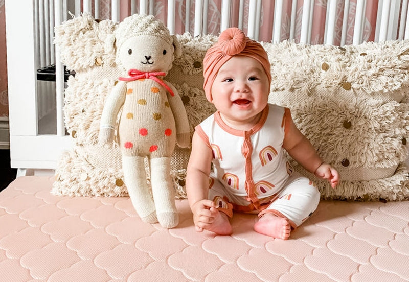 Happy baby sitting on a pink mattress with a stuffed bear, dressed in a white and orange patterned outfit and a matching headband, beside a crib with a fringed pillow.