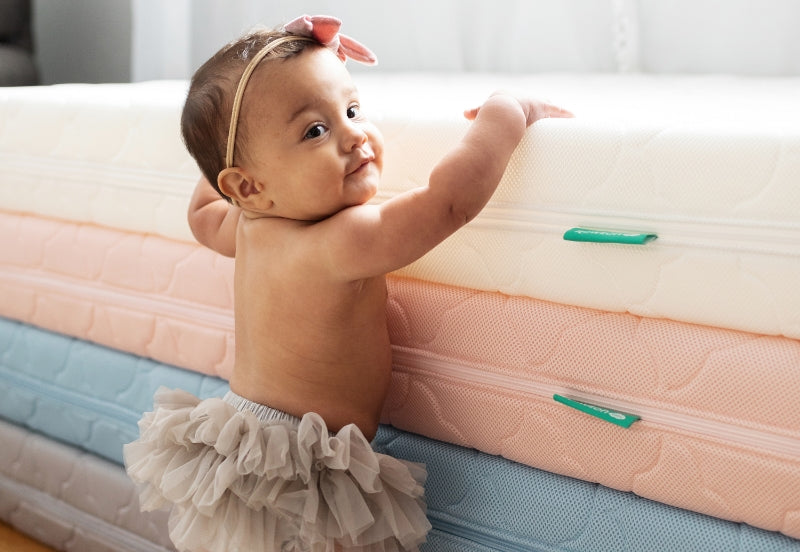 A baby in a beige ruffled skirt and headband smiles while standing next to a stack of colorful Newton Baby crib mattresses.