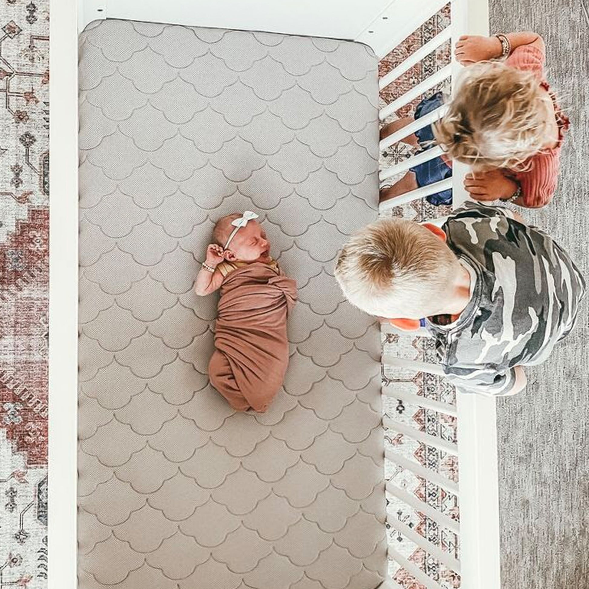 baby lying on newton grey crib mattress with siblings peering over crib guardrails at baby