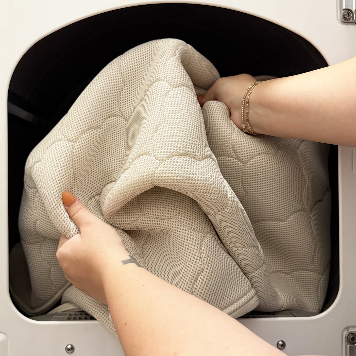 A person's hands place a washable, ultra-breathable Newton Baby Crib Mattress (in cream) into a front-loading washer—perfect for crib bedding.