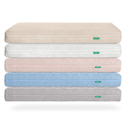A stack of five Newton Baby Crib Mattresses in beige, white, pink, blue, and gray is shown against a white background. Each mattress features ultra-breathable material, a subtle textured pattern, and a small green tag on the side.