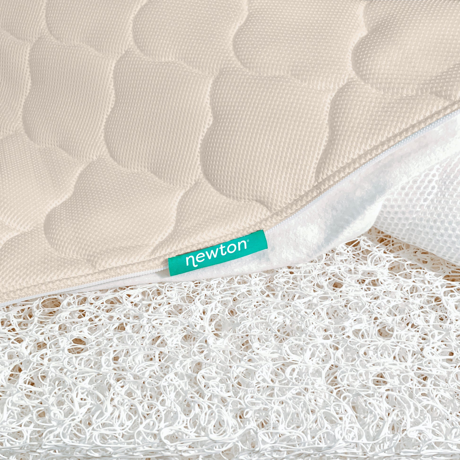 A close-up of a beige, quilted, washable Newton Baby Crib Mattress cover, partially unzipped to show its white ultra-breathable open-weave layer. A green “newton” tag is sewn into the seam.
