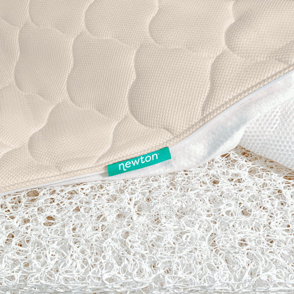 A close-up of a beige, quilted, washable Newton Baby Crib Mattress cover, partially unzipped to show its white ultra-breathable open-weave layer. A green “newton” tag is sewn into the seam.