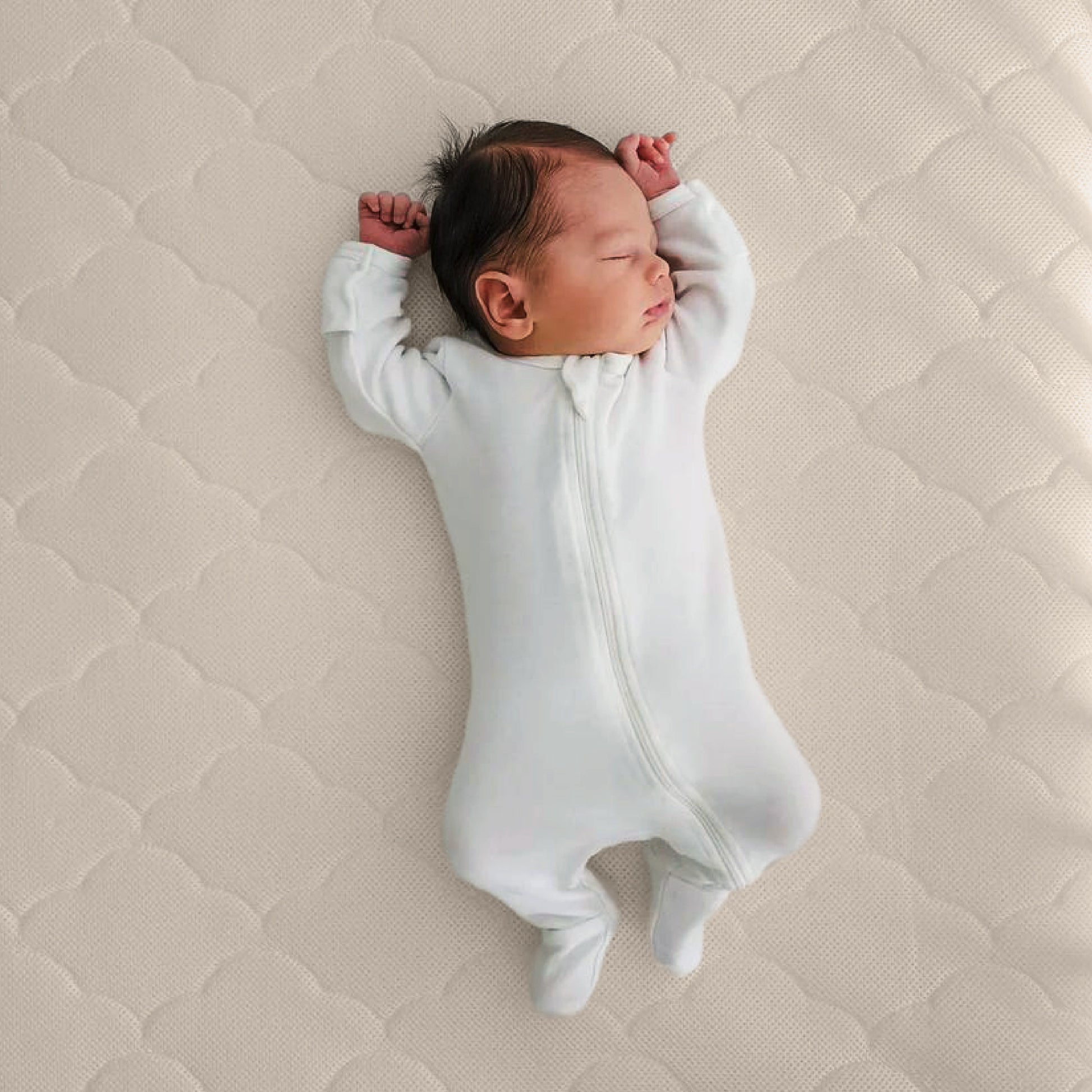 A newborn in a white zip-up onesie sleeps with arms raised on the Newton Baby Crib Mattress, which is ultra-breathable and light-colored.