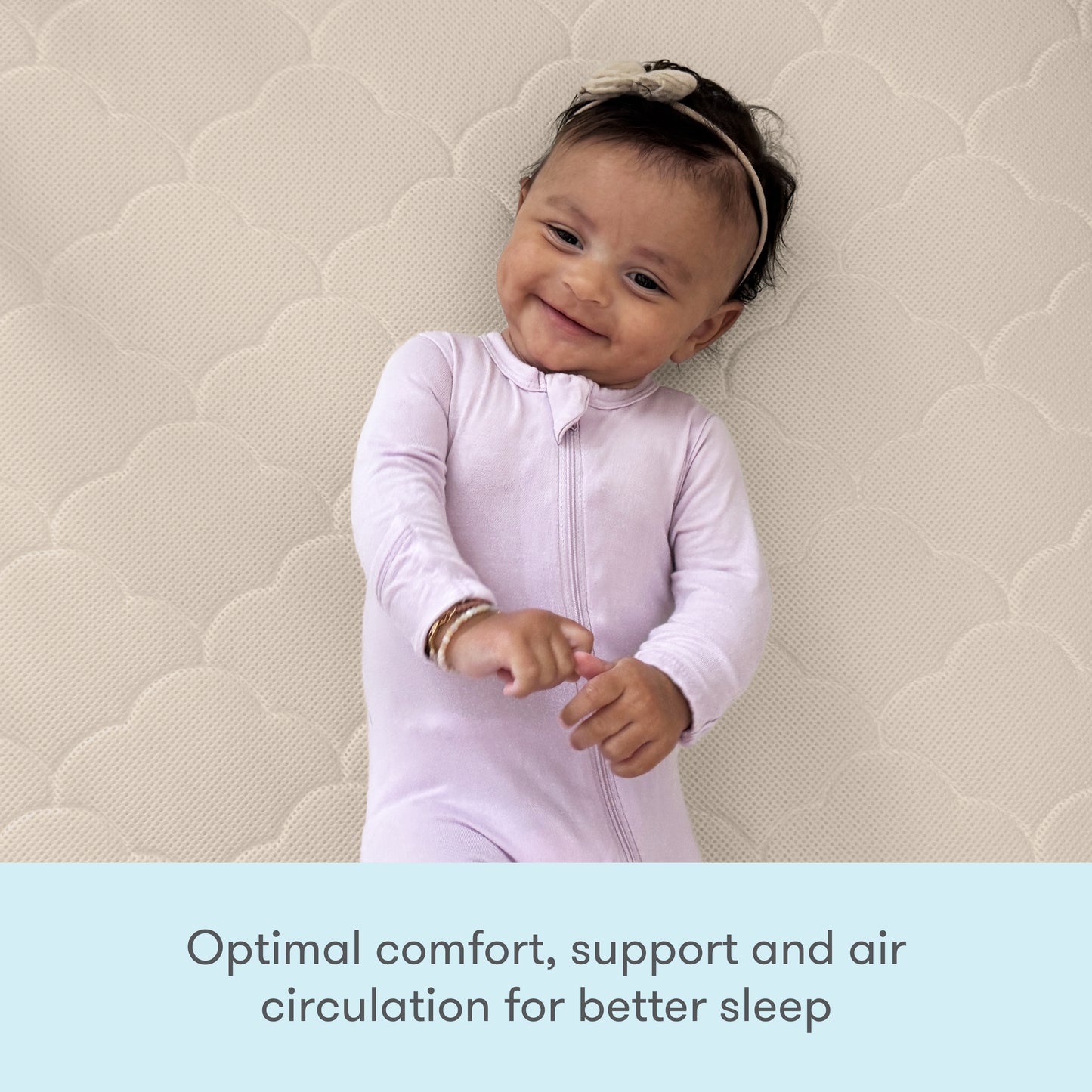 A happy baby in a light pink onesie rests on the Newton Baby Crib Mattress with a cloud pattern—offering optimal comfort, washable design, support, and enhanced air circulation for better sleep.