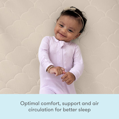 A happy baby in a light pink onesie rests on the Newton Baby Crib Mattress with a cloud pattern—offering optimal comfort, washable design, support, and enhanced air circulation for better sleep.