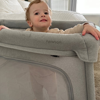 A toddler stands and smiles inside a light gray Newton playpen, holding onto the padded edge. A bed with white bedding and a window with blinds are visible in the background.