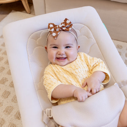 A smiling baby in a yellow outfit and a brown headband with white flowers sits indoors in the Newton Baby Rocker, holding the adjustable harness.