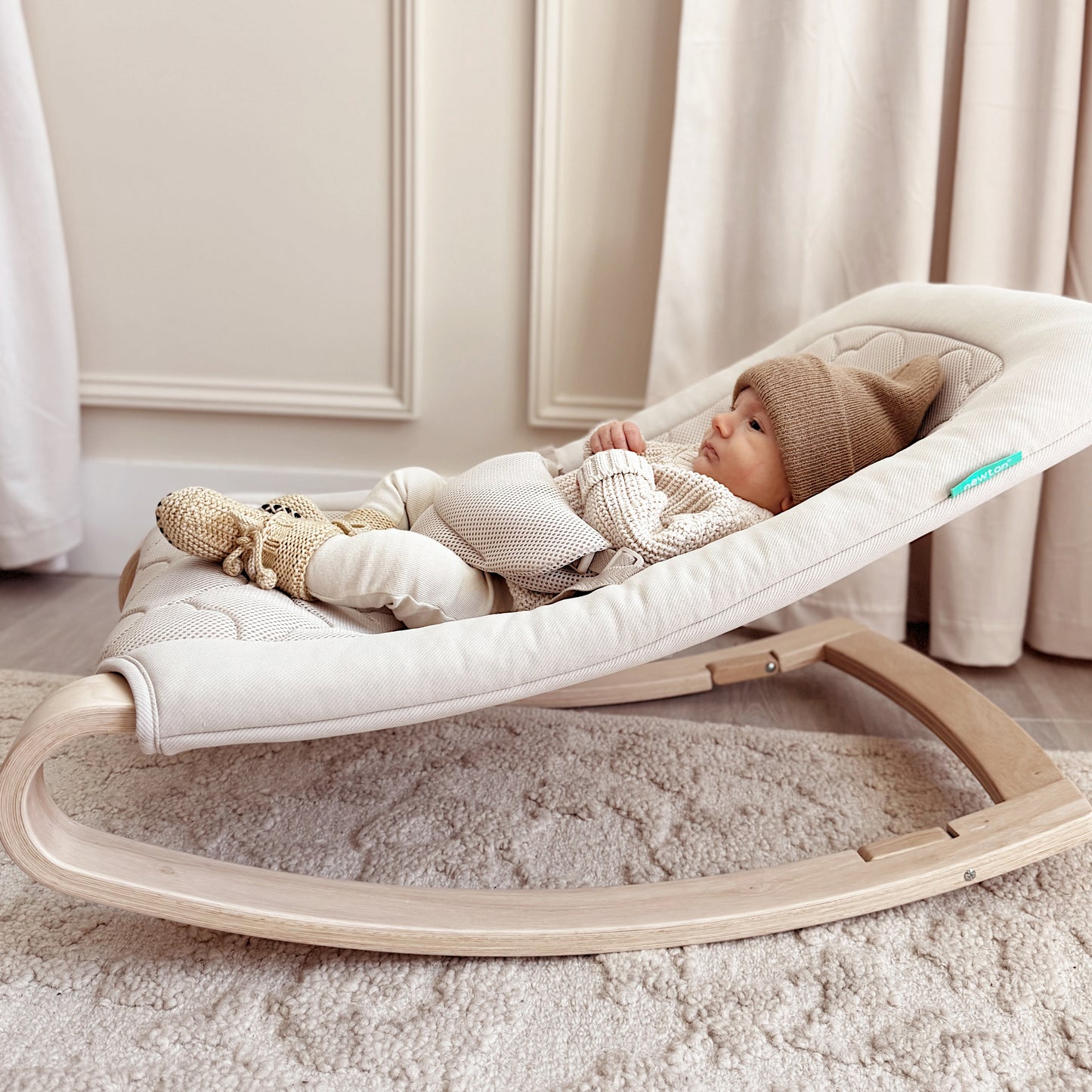 A baby in neutral tones lies in the Newton Baby Rocker, featuring a breathable seat cover and wooden frame, placed on a textured carpet in an elegantly lit room.