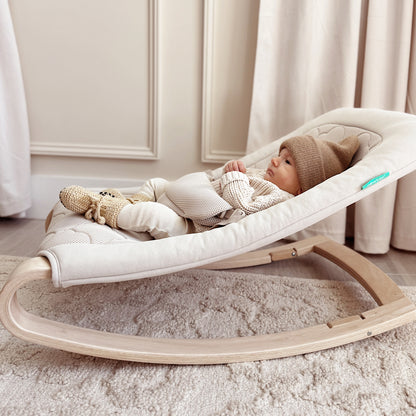 A baby in neutral tones lies in the Newton Baby Rocker, featuring a breathable seat cover and wooden frame, placed on a textured carpet in an elegantly lit room.