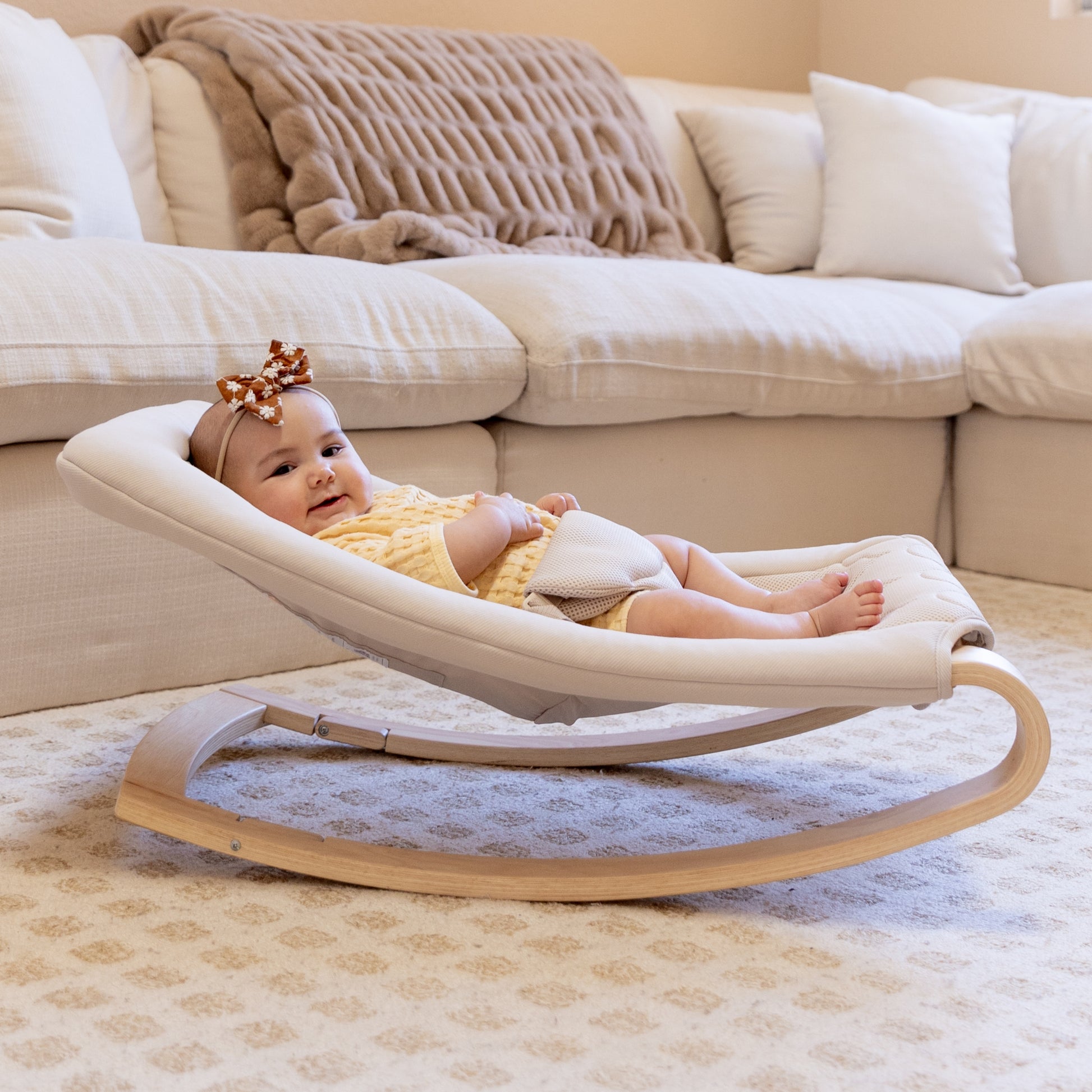 A baby in a yellow outfit and headband relaxes on the Newton Baby Rocker, which features a breathable seat cover, in a cozy living room with a cream sofa and patterned rug.