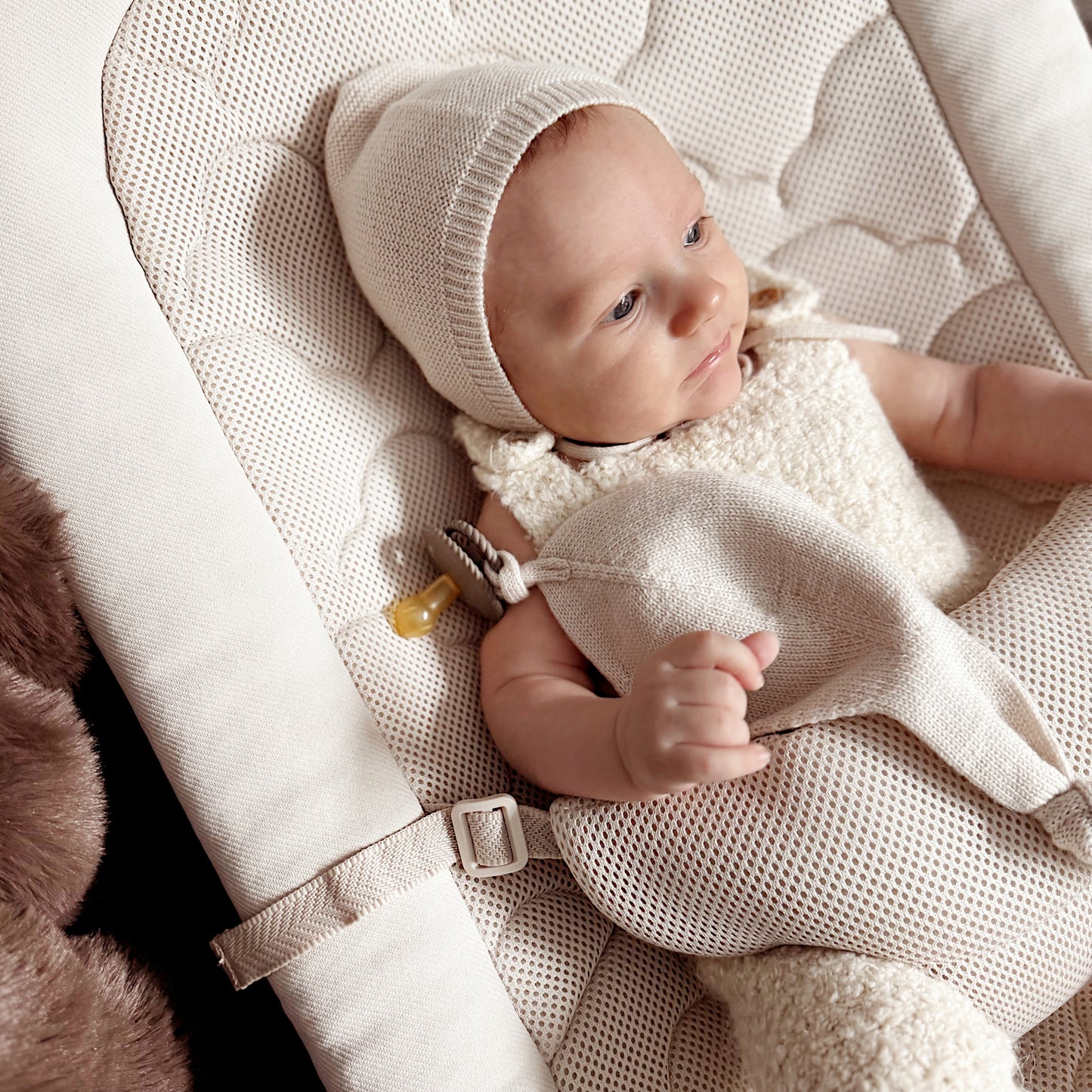 A baby in a cream outfit and bonnet lies on the Newton Baby Rocker, featuring a breathable seat cover, holding a soft blanket with a pacifier clipped to the side.