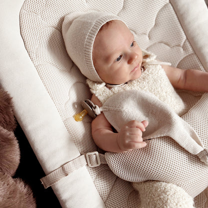 A baby in a cream outfit and bonnet lies on the Newton Baby Rocker, featuring a breathable seat cover, holding a soft blanket with a pacifier clipped to the side.