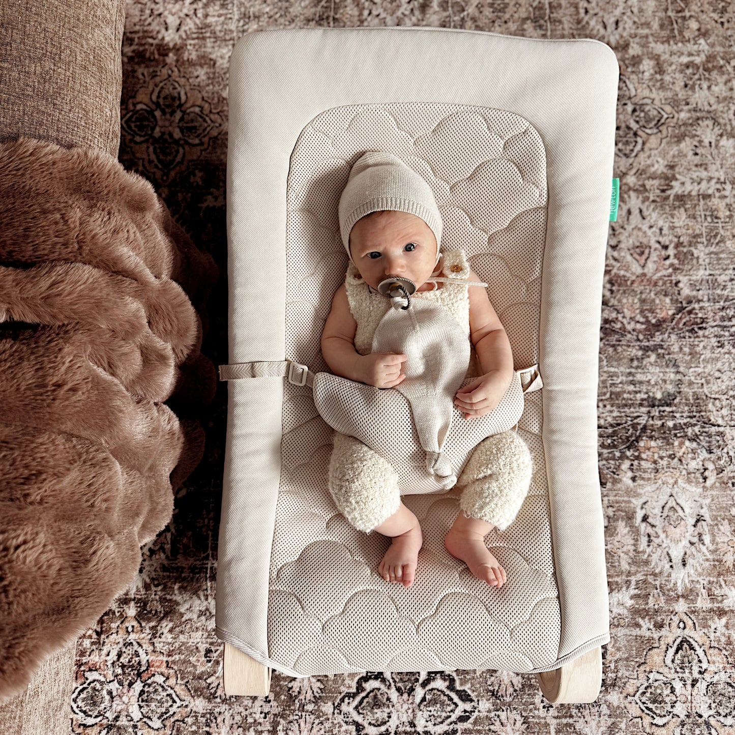 A baby in a knit hat and light outfit lies in the Newton Baby Baby Rocker, which features an adjustable harness, with a pacifier, on a patterned rug next to a brown furry blanket.