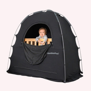 baby sitting in crib, underneath black slumberpod blackout tent skip