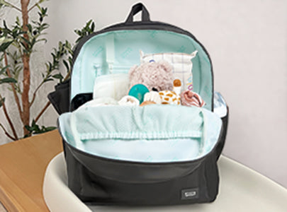 A black backpack sits open on a table, revealing neatly packed baby items including a stuffed animal, a pacifier, a bottle, and soft blankets, all arranged against a light blue interior.