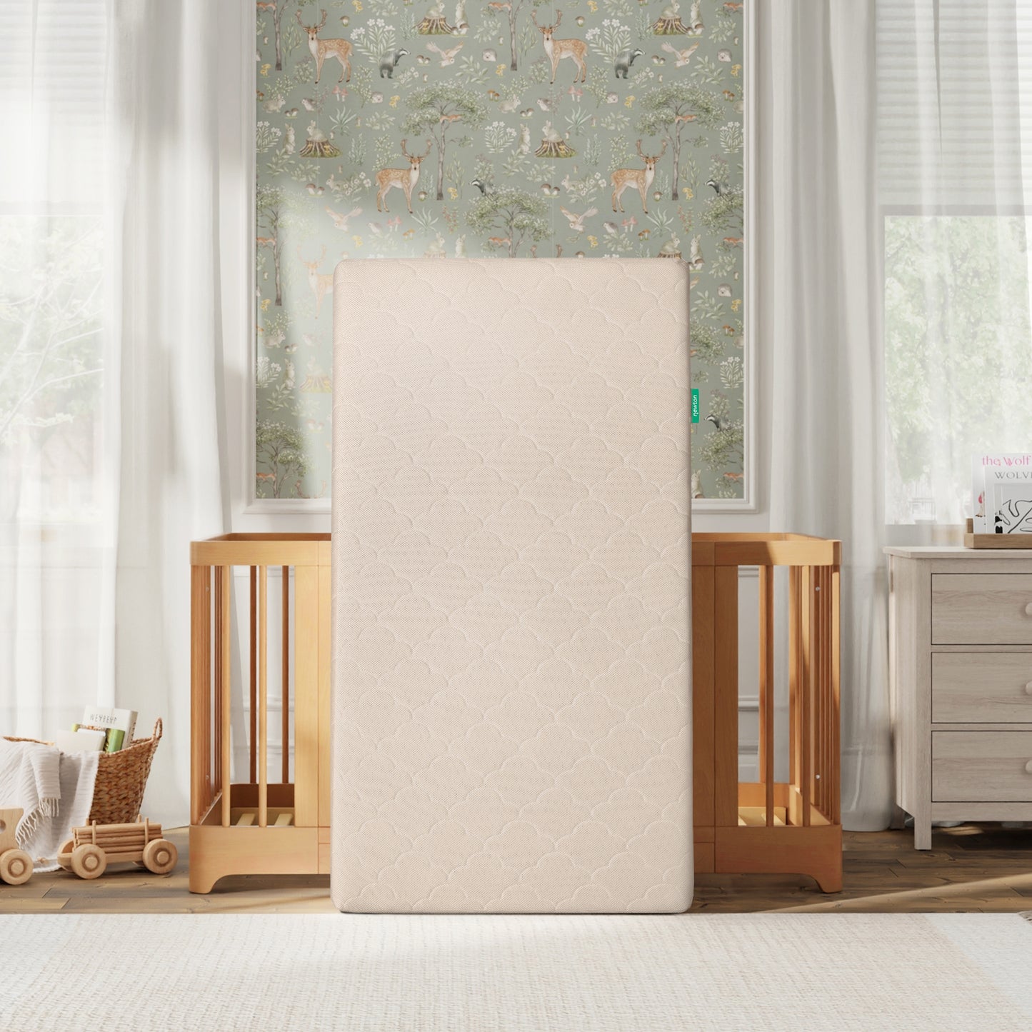 The Newton Baby Crib Mattress, made to be breathable and washable, stands upright in front of a wooden crib in a softly lit nursery with animal wallpaper, white curtains, a dresser, and a toy wagon on the wooden floor.