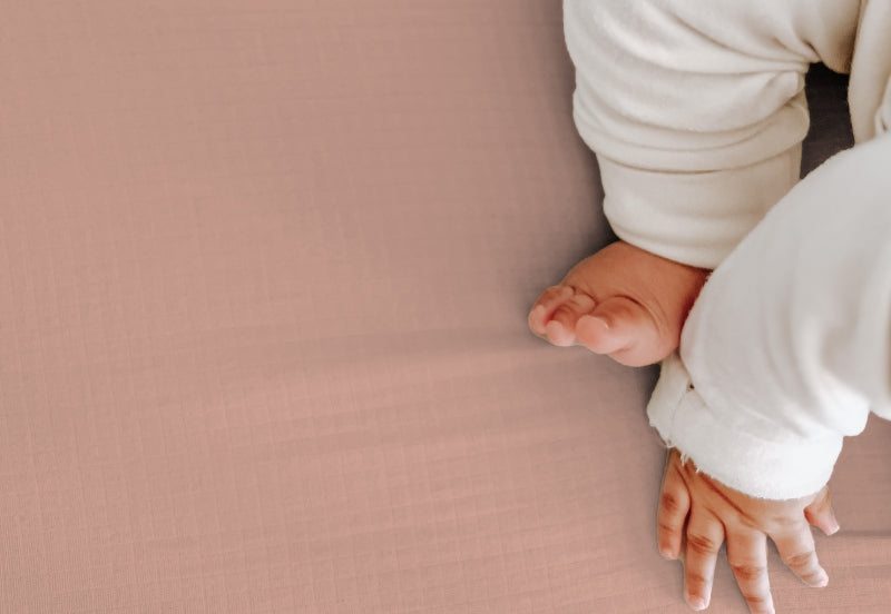 Close-up image of a baby's bare feet and hands on a soft pink surface, dressed in a white outfit. 999-3020-CLY 999-3520-CLY