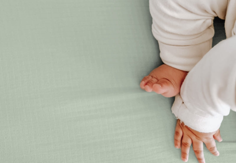 A close-up image of a baby's little feet and hands on a soft sage colored sheet. 999-3020-SNS 999-3520-SNS