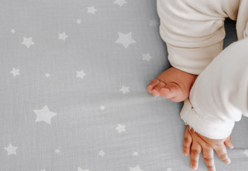 A close-up of a baby's feet and hands on a gray sheet with a white star pattern. 999-3020-SGW 999-3520-SGW