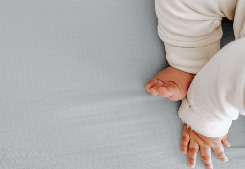 Close-up of a baby's hand and foot on a textured twilight grey sheet, conveying softness and comfort. 999-3020-GNG 999-3520-GNG
