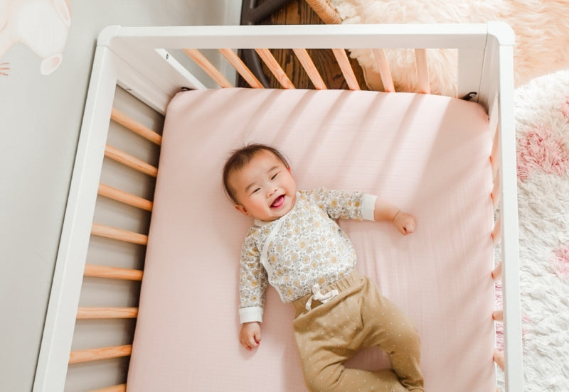 A joyful baby lying on a pink mattress in a white crib, smiling broadly and dressed in a floral shirt and tan pants. all