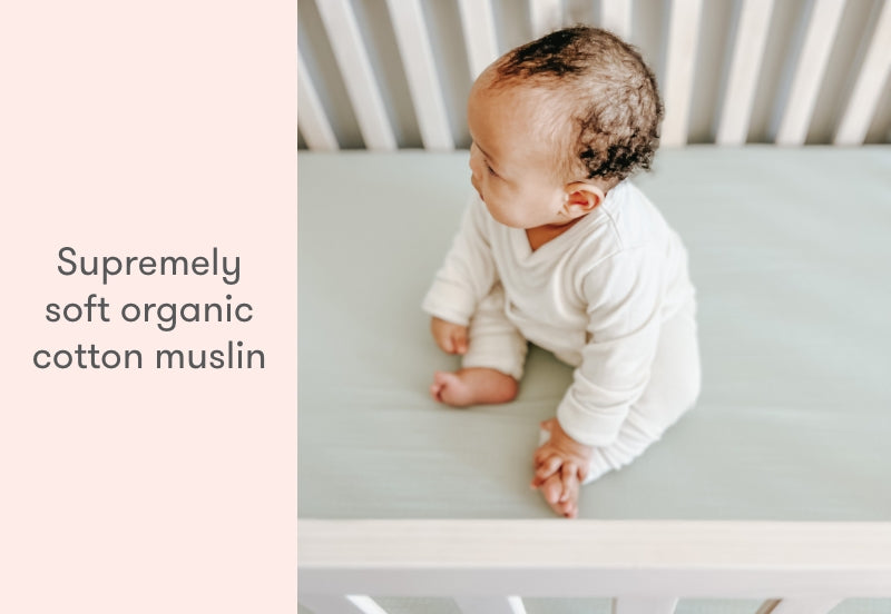 Baby sitting on a crib mattress covered with a supremely soft organic cotton muslin sheet. all
