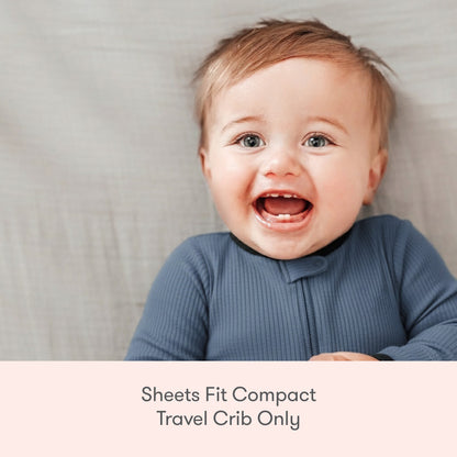 A smiling baby in a blue onesie lies on a light gray sheet, with text below reading: Newton Baby Compact Travel Crib Sheets fit compact travel cribs only.