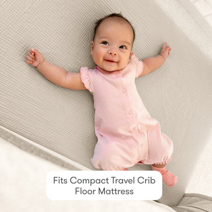 A smiling baby in a pink striped outfit lies on a light quilted mattress with Newton Baby Compact Travel Crib Sheets. Text below reads: Fits Compact Travel Crib Floor Mattress.