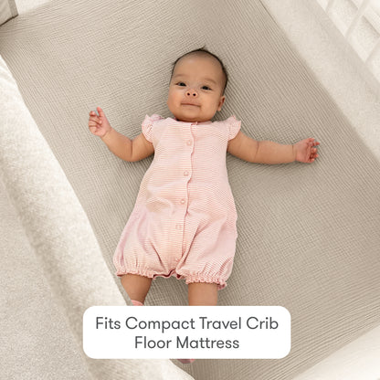 A baby in a pink romper lies on a light gray mattress in a crib. Text reads, "Newton Baby Compact Travel Crib Sheets: snug fit for added comfort on your travel crib mattress.