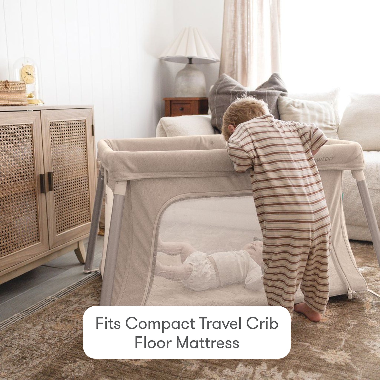 A toddler in striped pajamas leans over a beige travel crib to look at a baby lying on Newton Baby Compact Travel Crib Sheets. Cozy living room with sofa, lamp, and cabinet. Text: Fits Compact Travel Crib Floor Mattress.
