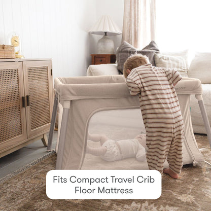 A toddler in striped pajamas leans over a beige travel crib to look at a baby lying on Newton Baby Compact Travel Crib Sheets. Cozy living room with sofa, lamp, and cabinet. Text: Fits Compact Travel Crib Floor Mattress.