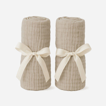 Two beige, rolled-up blankets stand upright, each tied with a cream ribbon, perfectly complementing your Newton Baby Compact Travel Crib Sheets—shown against a plain white background.